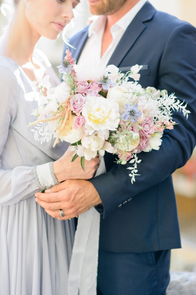 Bouquet de pivoines et fleurs pastel tenu par la mariée en robe bleu-gris, le marié en costume bleu