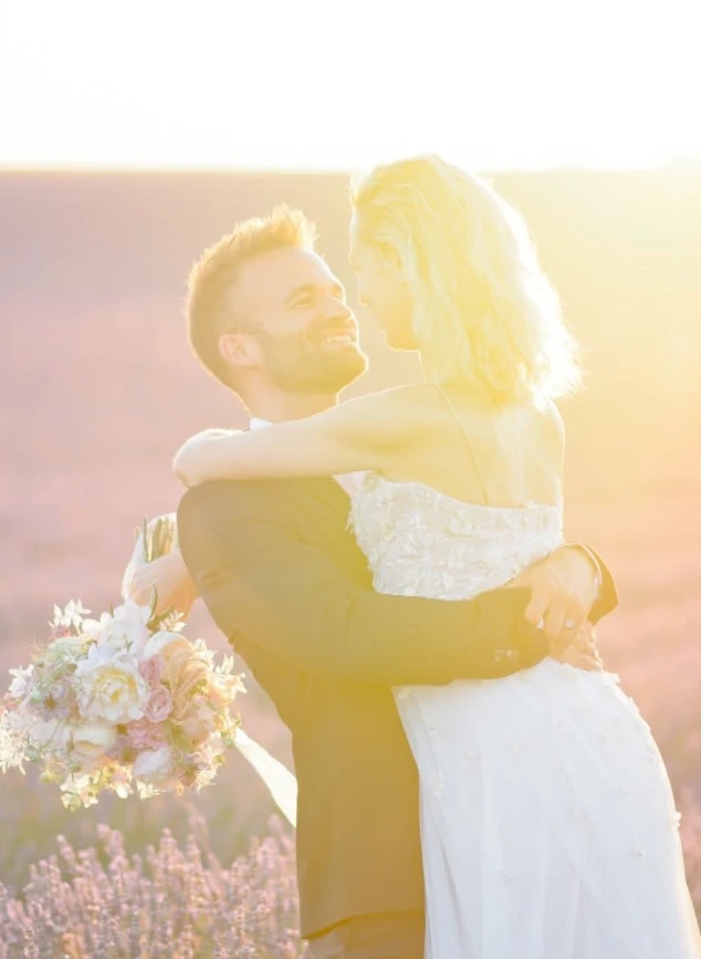 Le marié soulève sa femme dans un champ de lavande à contre-jour, tous deux enlacés et souriants, le bouquet à la main
