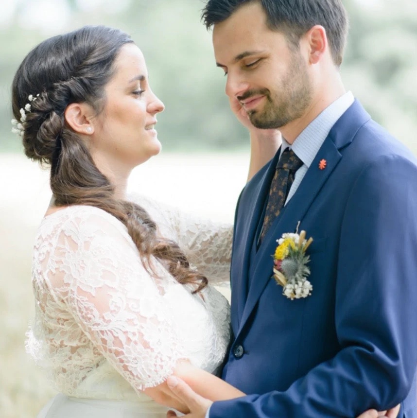 Les mariés face à face, front contre front, la mariée en robe de dentelle et le marié en costume bleu à boutonnière champêtre