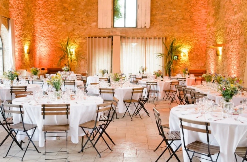 Salle de réception aux murs de pierre ocre, tables rondes dressées de blanc et bouquets champêtres