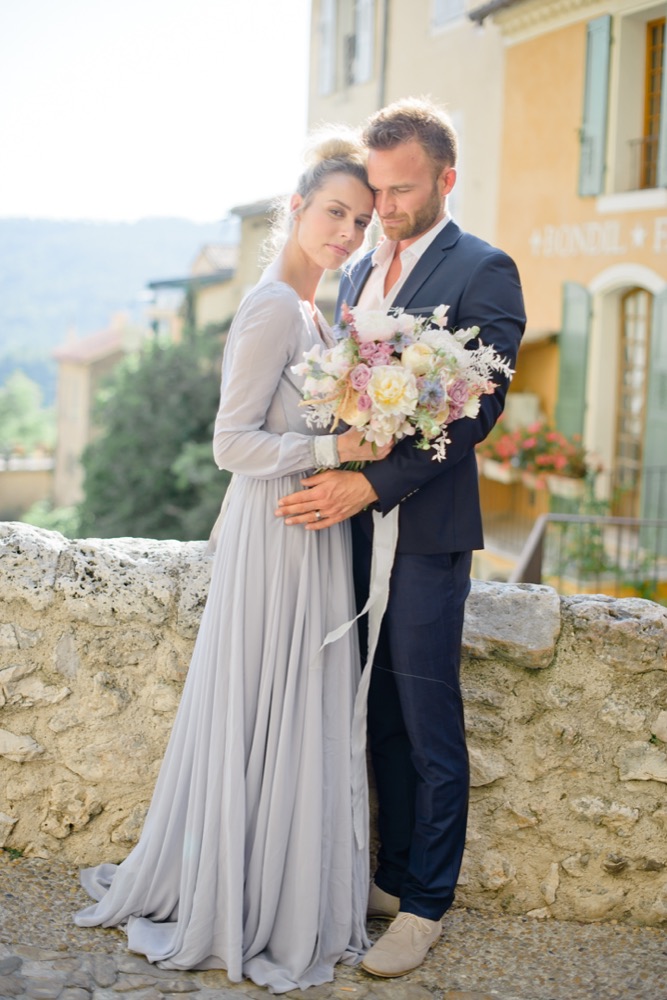 Couple enlacé avec un bouquet champêtre devant les façades ocre d'un village